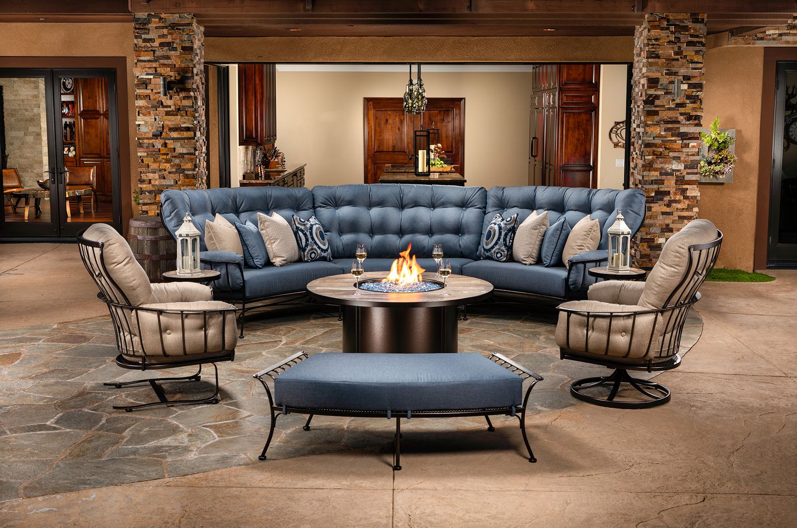 A curved wrought-iron sectional with blue cushions gathered around a fire table under a covered patio