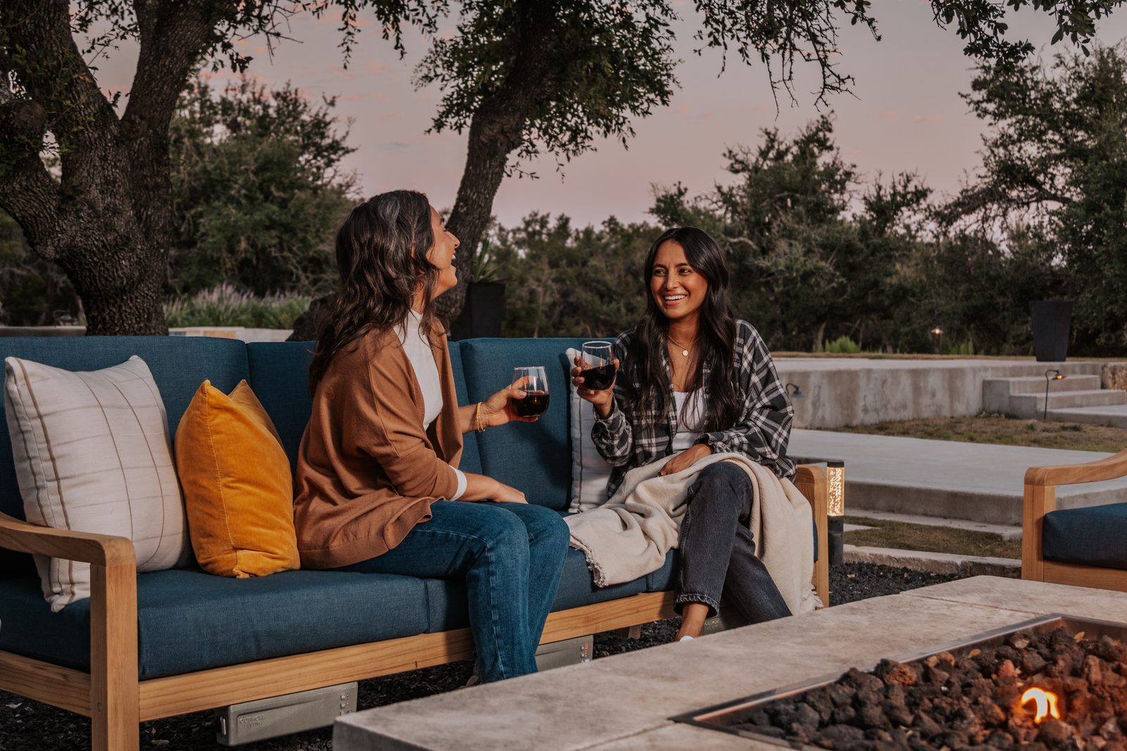 Two friends with drinks on a teak deep-seating sofa beside a fire pit at dusk